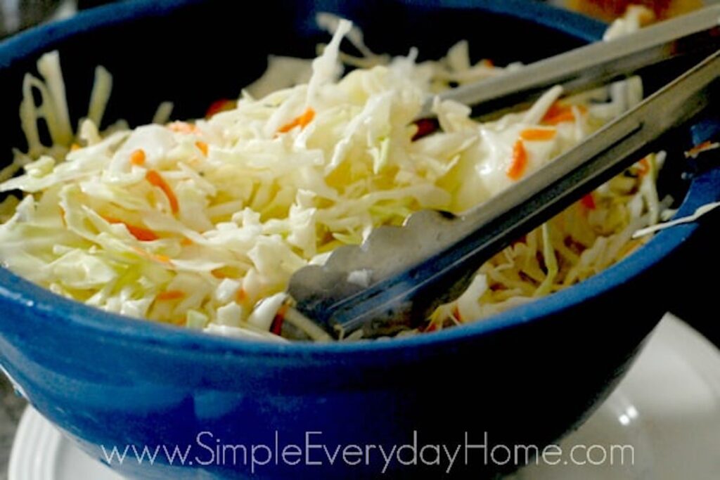 Cut cabbage and shredded carrot being tossed in blue colander with metal tongs