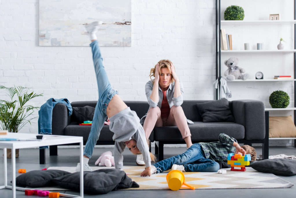 A woman sitting on the sofa looking overwhelmed while her kids play and do cartwheels in the living room