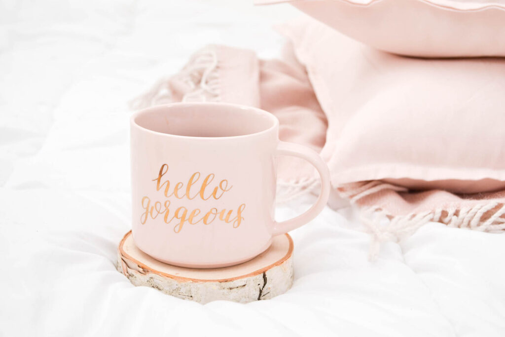 a mug with the words "hello gorgeous" beside pink pillows on a bed