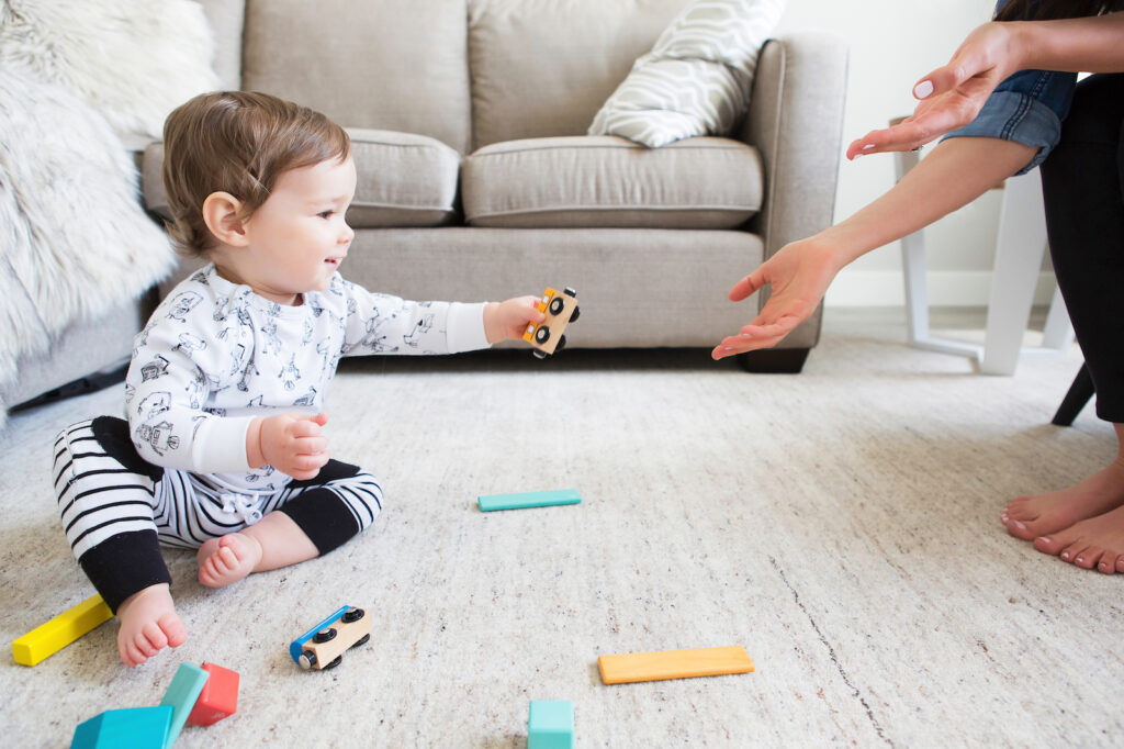 A baby sitting on the floor playing with wooden trains and handing a piece to his mother