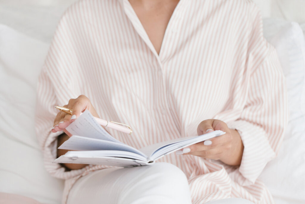 Woman in pink striped shirt sitting on bed and writing in notebook