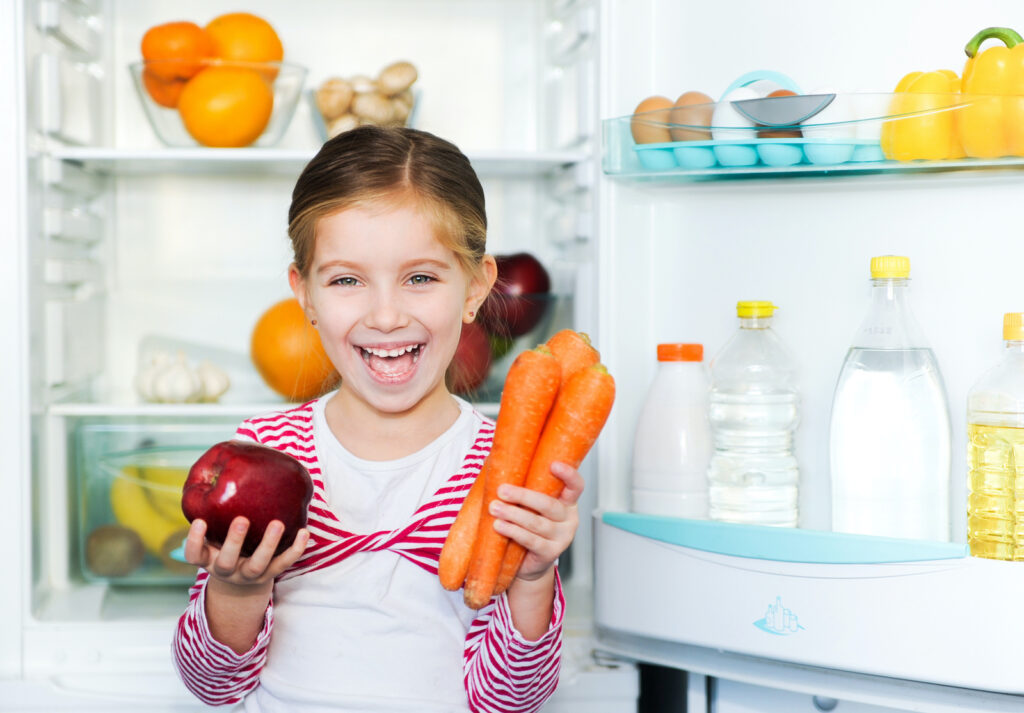 A little girl holding an apple and some carrots and smiling in front of an open refrigerator