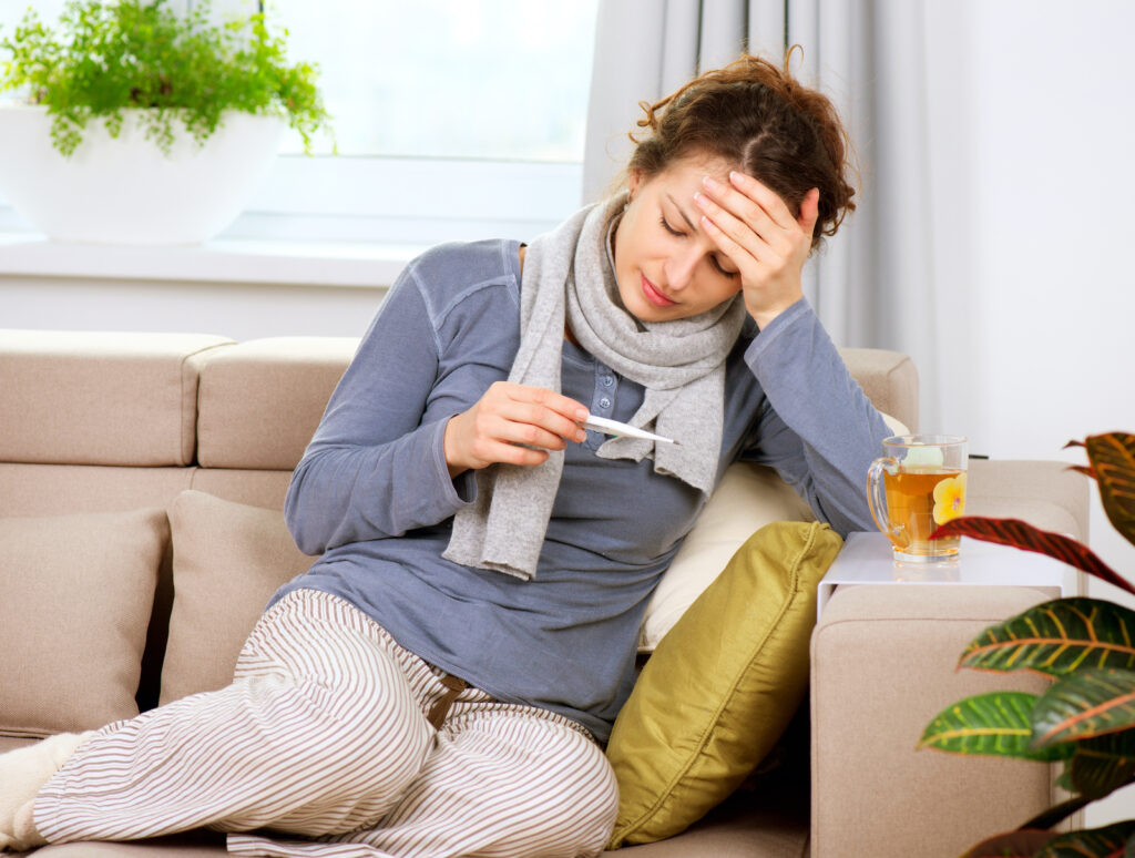 A woman on a sofa holding her head and looking at a thermometer